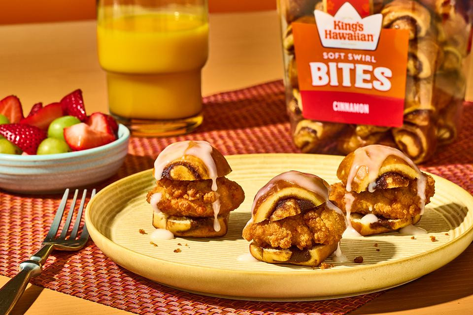 Three mini chicken sliders made with King's Hawaiian Soft Swirl Cinnamon Bites, crispy fried chicken, and vanilla icing drizzle, served on a plate with strawberries, green grapes, and orange juice.