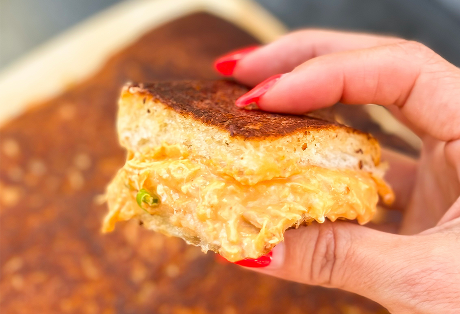 A hand holding a halved Buffalo Chicken Dip Grilled Cheese made with King's Hawaiian Sweet Rolls, showing a gooey, cheesy buffalo chicken filling with green onions and a golden-grilled crust, held over a baking dish of buffalo chicken dip.