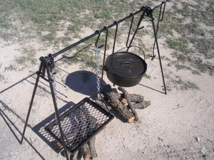 A high-angle bird's-eye view looking down at the steel cooking frame. This perspective highlights the sturdy construction of the A-frame supports, the chain-link suspension for the grill, and the various sizes of "S" hooks used to hang cookware over the fire.