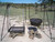 A front-facing, eye-level shot of a portable campfire cooking rig in an open field. The black metal frame holds a hanging mesh grill and a lidded Dutch oven at different heights, demonstrating the adjustable hooks and chains used for heat control over two separate wood piles.