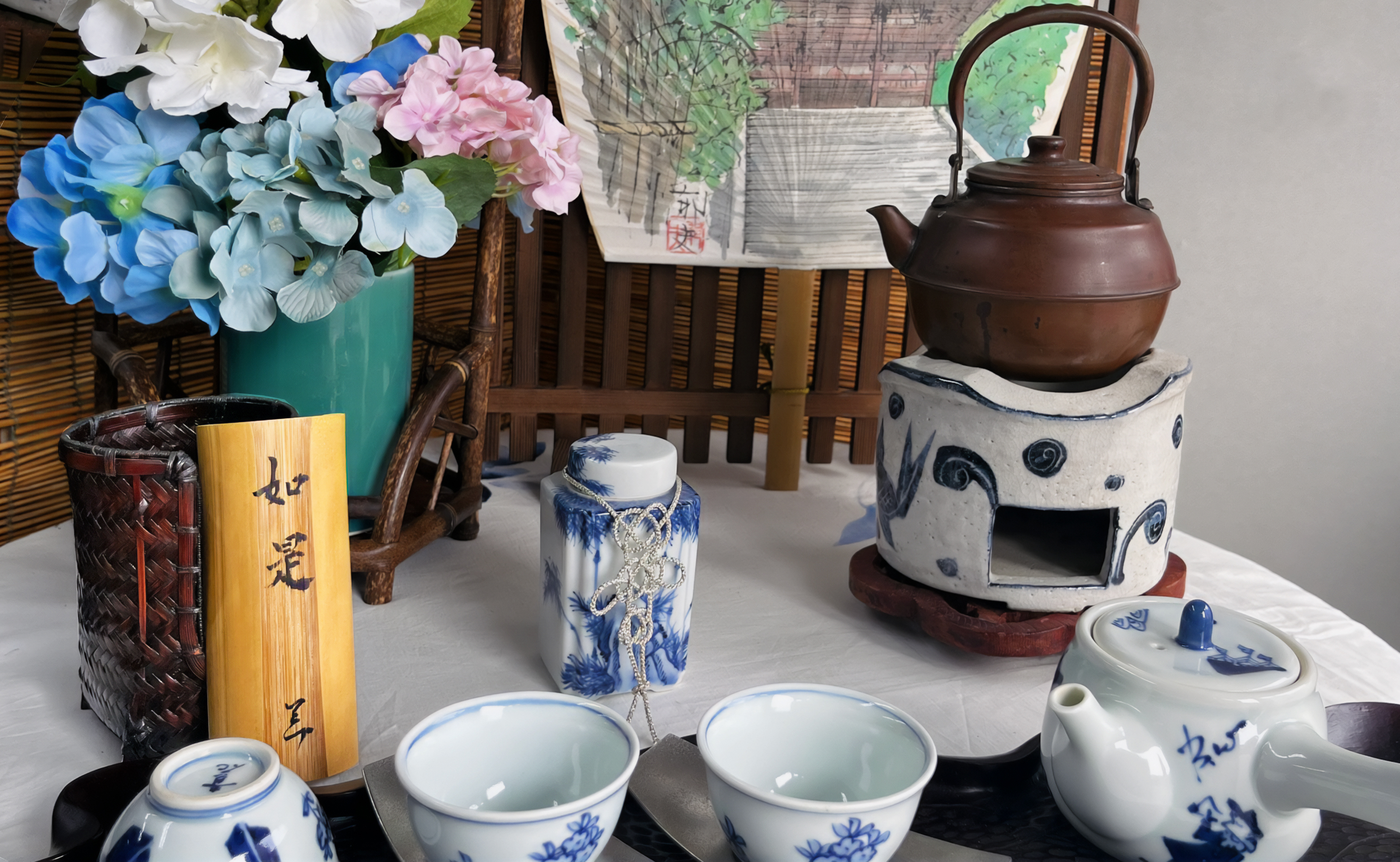 Early summer Japanese tea ceremony display with iron kettle, porcelain tea set, bamboo calligraphy, and hydrangeas arranged around a hand-painted brazier and a garden gate painting backdrop