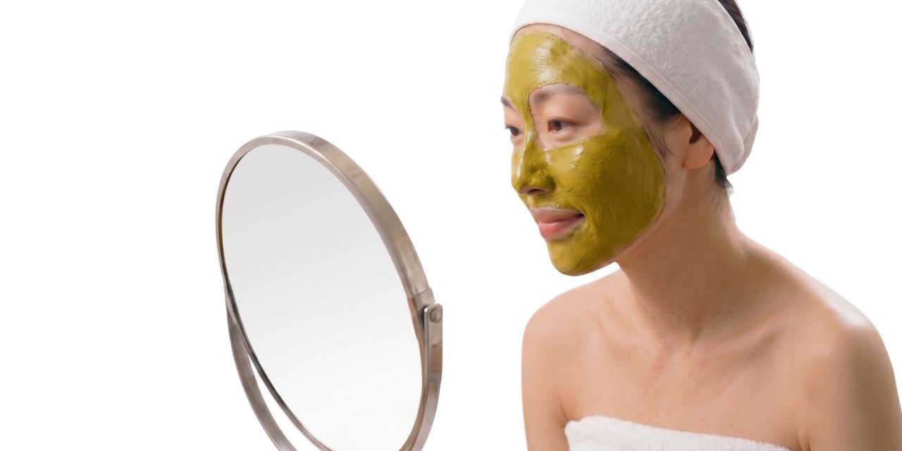 Japanese woman wearing a traditional green facial mask and towel headband, looking into a mirror during a relaxing at-home skincare treatment for hydration, brightening, and skin renewal.