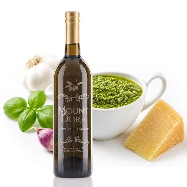 A tall dark-glass bottle of Mount Dora Olive Oil Company with a gold screw cap stands on a white surface. Behind it sits a white ceramic bowl of bright green pesto, with fresh basil leaves, a garlic bulb, a purple shallot, and a wedge of aged Parmigiano-Reggiano arranged around the bottle.