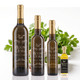 Four dark glass bottles of Mount Dora Olive Oil Company Chimichurri Verde olive oil in descending size, 750 ml, 375 ml, 200 ml, and 60 ml, each with a gold cap. Fresh parsley, garlic, a sliced green chili, and a bowl of chimichurri sauce sit behind them on a white background.