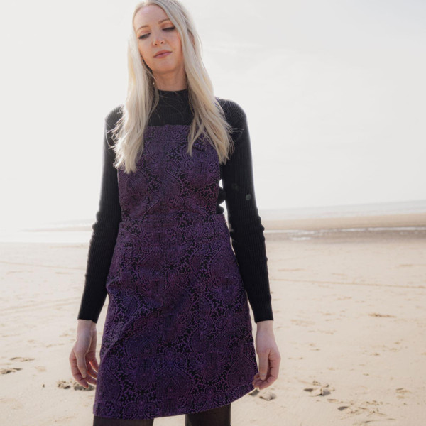 Madcap England Retro 70s Paisley Twill Dungaree Dress in Black and Purple MC1113. Woman weaing Madcap England Pinafore Dress in Purple