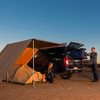 ARB 4X4 Accessories Expedition Awning Front Wind Break 2.5m ARB, outdoor camping setup with vehicle and tents at sunset, showcasing the awning installed on a vehicle with wind break for camping comfort.