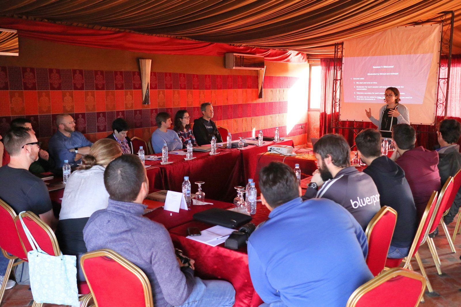 Bluespark team members seated around a U-shaped table in a red-draped tented room in Morocco, listening to a presenter standing beside a projected slide titled Session 1: Welcome.
