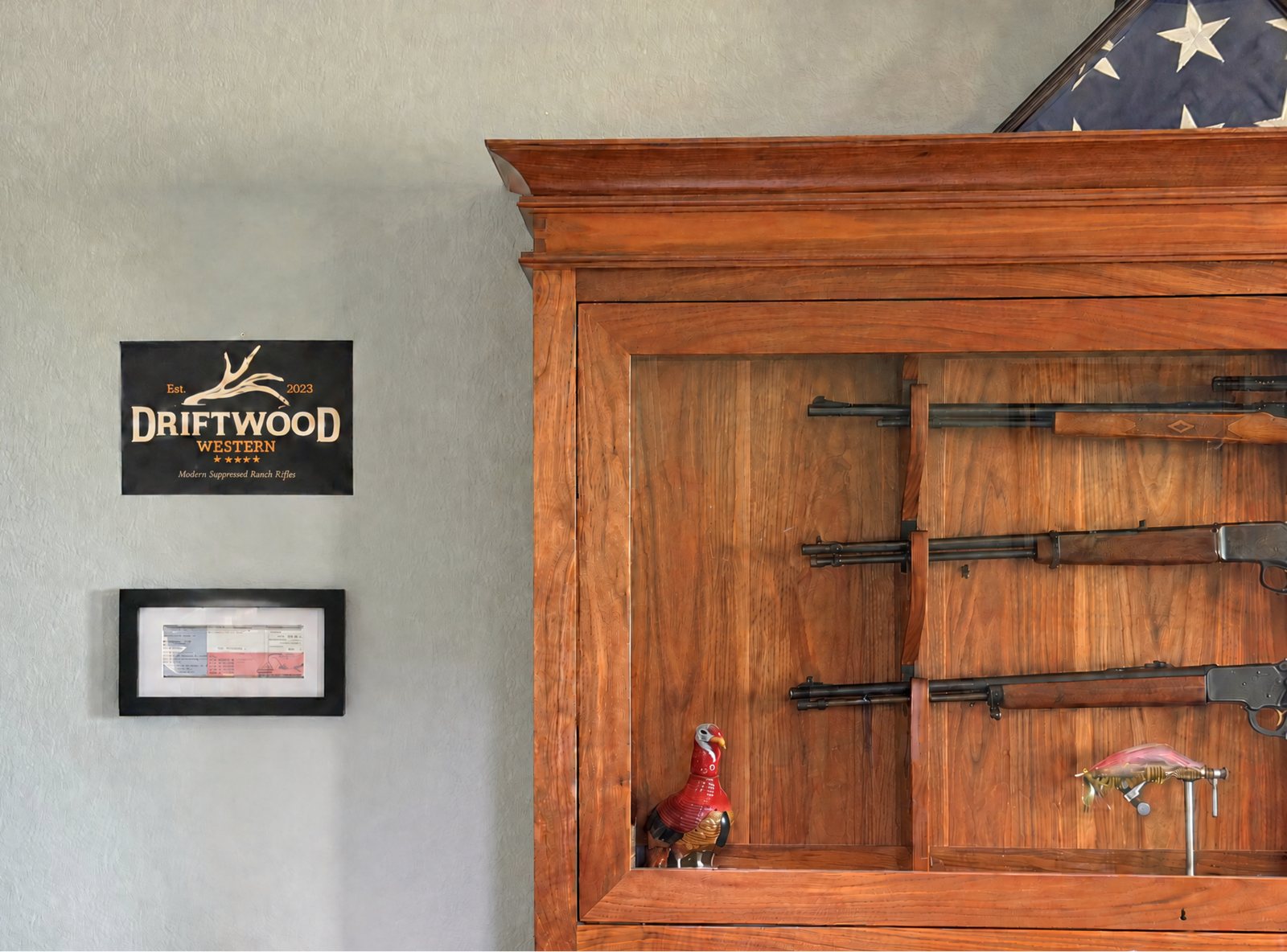A wooden gun cabinet with a glass front in the Driftwood Western office, holding three family rifles, beside a framed Driftwood Western sign on a gray plaster wall, with a folded flag resting on top.
