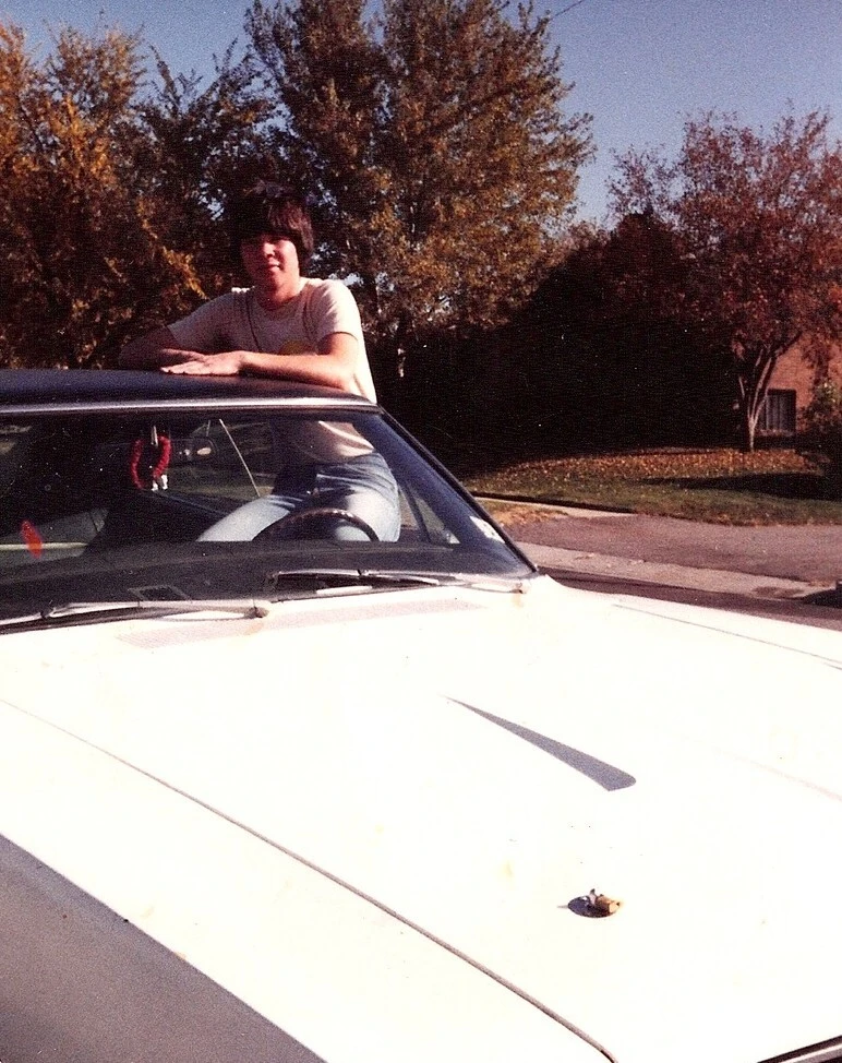 Archival color snapshot of Michael Tucker at about sixteen, standing behind a white car and resting his arms on the roof, with autumn trees along a residential street behind him.