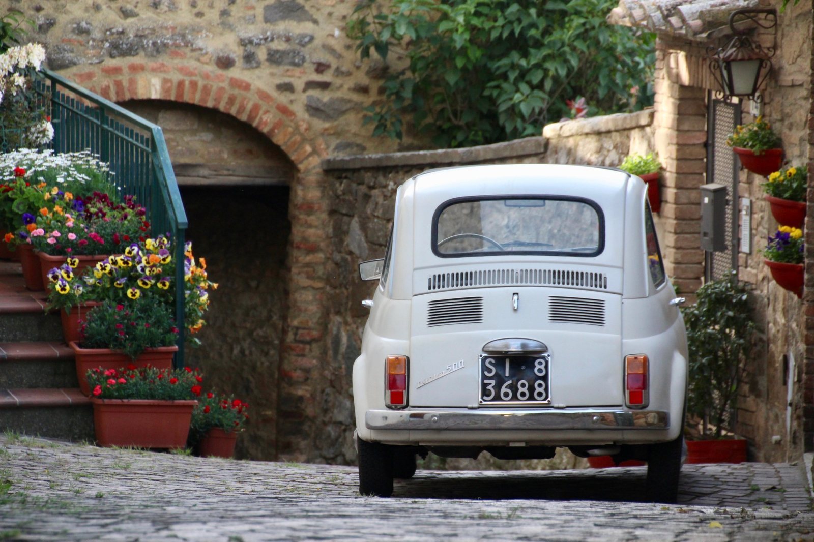 A white vintage Fiat 500 parked on a cobbled lane between stone walls in an Italian village, beside steps lined with flowerpots.