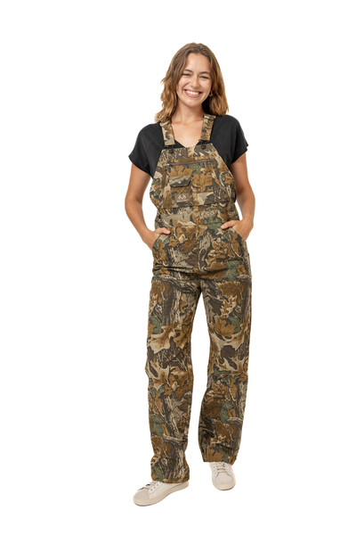 Woman wearing Realtree Advantage Classic canvas hunting bib overalls, shown in a full-body front view with hands in pockets.