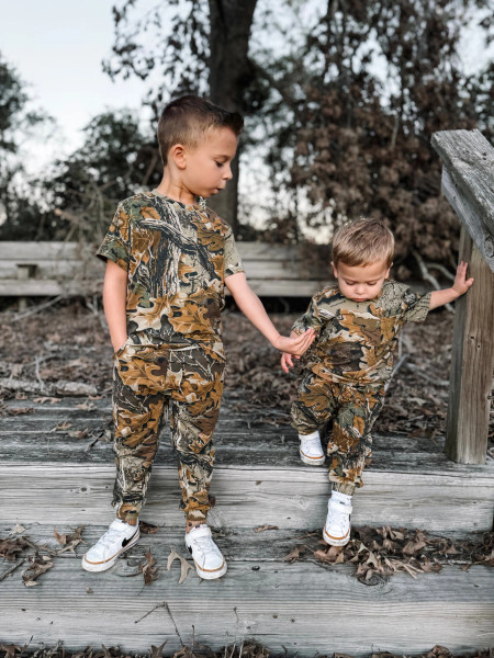 Two young boys wearing Realtree Advantage Classic camo sweatpants outdoors on wooden steps surrounded by fallen leaves.