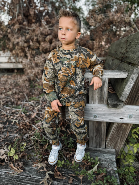 Child wearing Realtree Advantage Classic camo sweatpants and sweatshirt leaning against wooden railing in wooded setting.