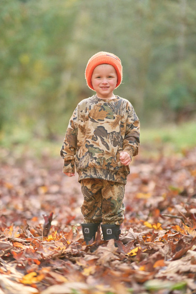 Toddler in Realtree Advantage Classic camo sweatshirt and joggers walking through fall leaves with an orange beanie.