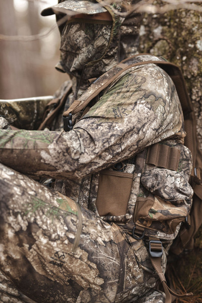 Side view of hunter seated against tree wearing Realtree APX turkey vest with accessible storage pockets.