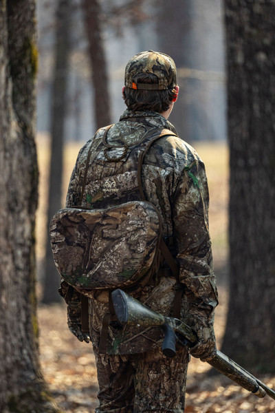 Rear view of hunter wearing Realtree APX turkey vest with attached seat and shotgun in wooded setting.