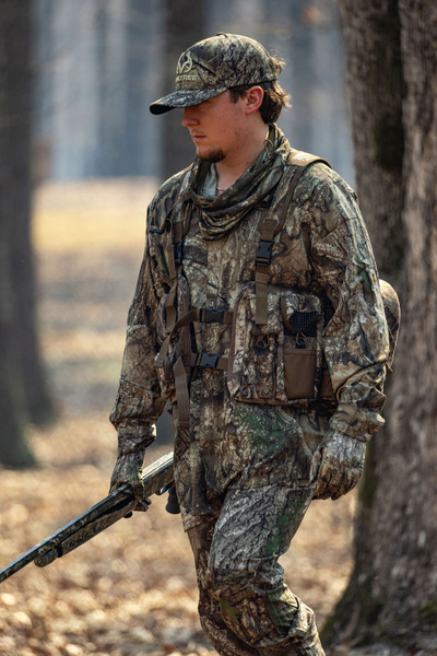 Hunter walking through woods wearing Realtree APX turkey vest with front storage pockets and shotgun.