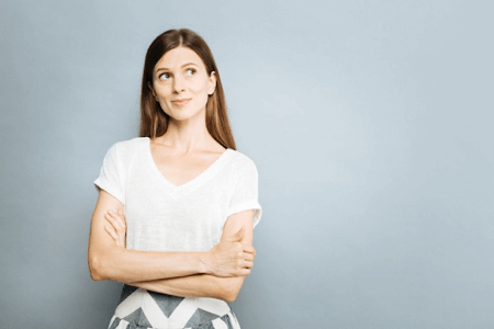 Woman in white shirt against gray background with crossed arms looking up in thought