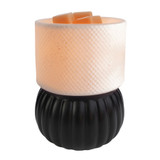 A tabletop Luxe Lamp Illumination Fragrance Warmer featuring a matte black ribbed base and a glowing white textured ceramic shade filled with wax melts