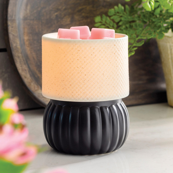 A close-up of the Luxe Lamp Illumination Fragrance Warmer on a white marble surface, showing the warm glow through the diamond-textured shade and pink wax melts on top, styled with green foliage