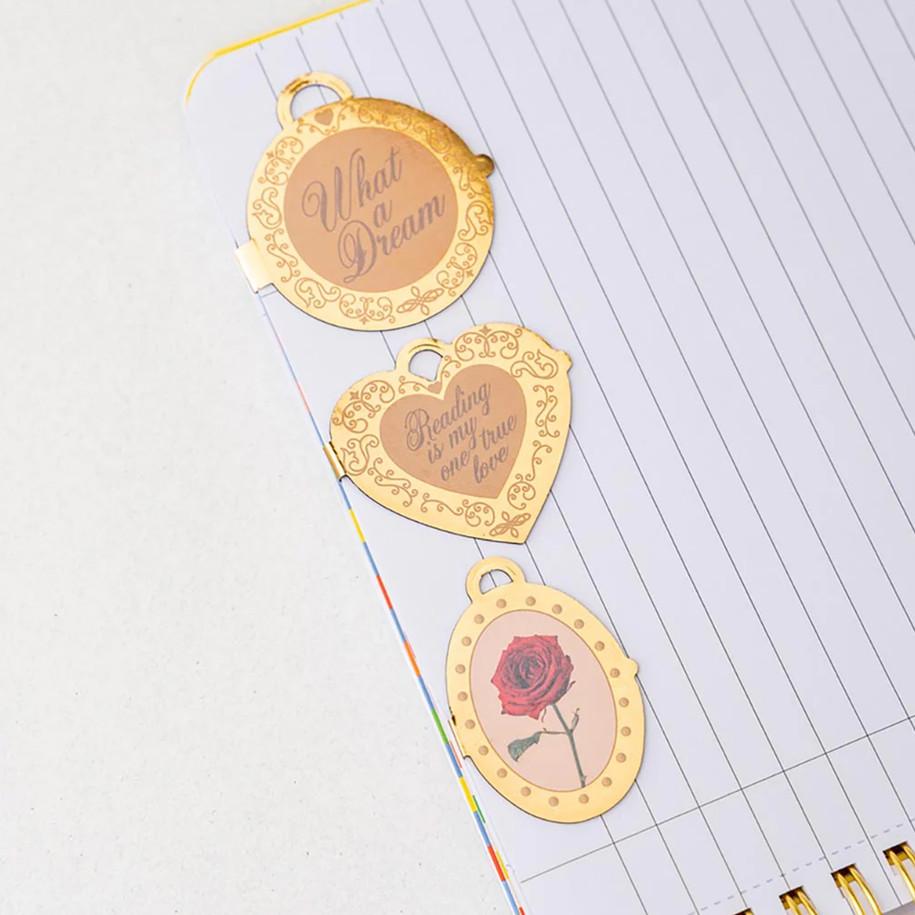 Three locket-style magnetic bookmarks featuring a rose and romantic text clipped to the side of a lined notebook page.