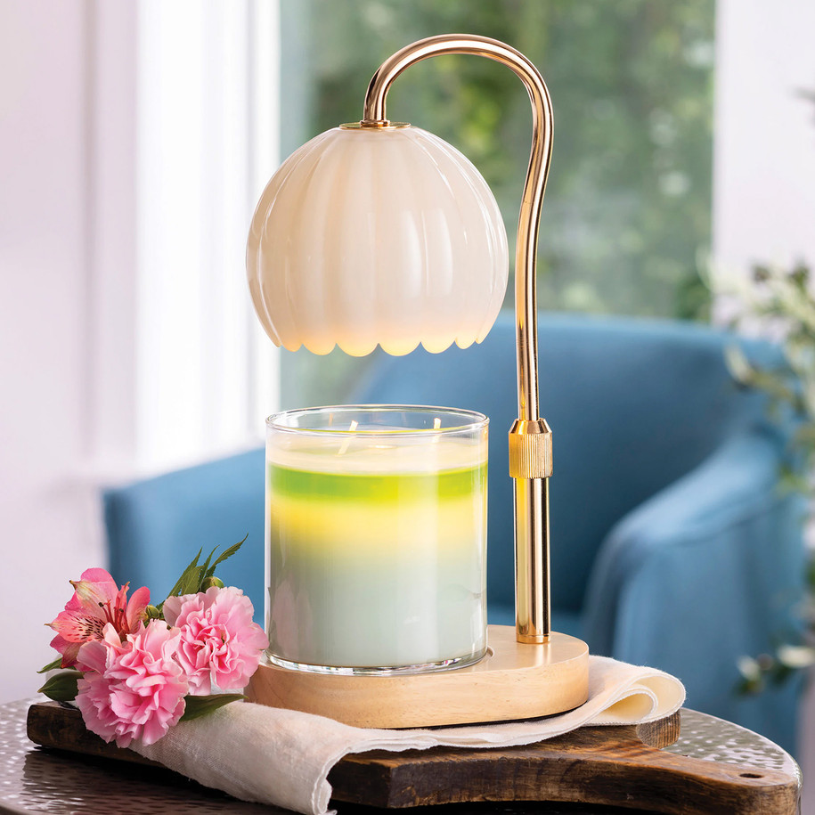 A close-up of the Milk Glass Candle Warmer Lamp in use, showing the warm amber glow through the scalloped shade as it melts a jar candle next to fresh pink flowers