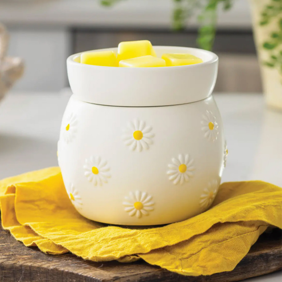 The Daisy Wax Warmer sitting on a bright yellow cloth and wooden board, warming yellow wax melts to create a cheerful, spring-inspired atmosphere