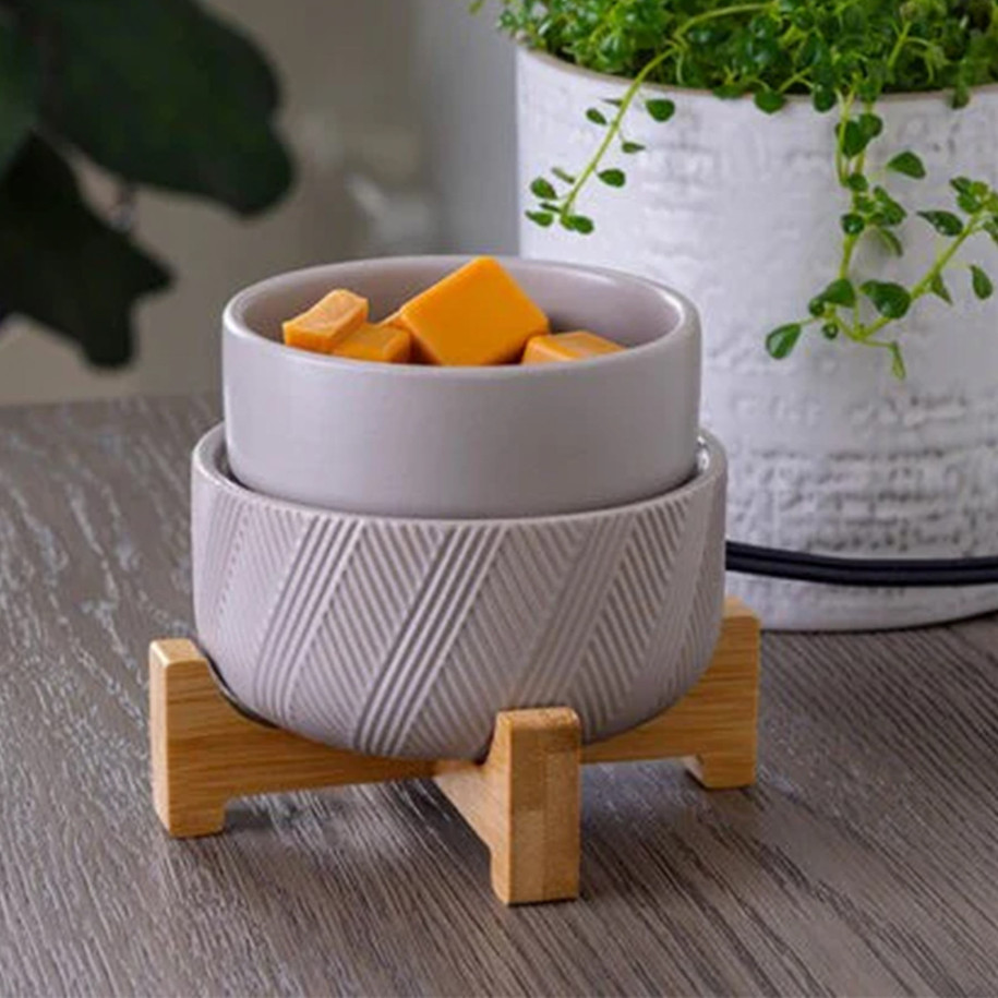 A close-up of the grey textured 2-in-1 warmer in use with the ceramic dish, filled with golden-brown wax cubes and positioned on a wooden surface next to a white potted plant
