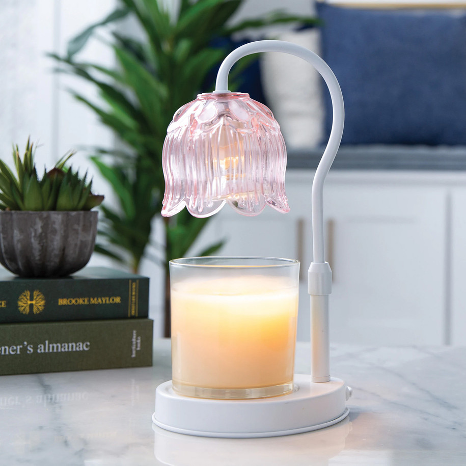 A pink Tulip Candle Warmer Lamp illuminating a large jar candle on a marble tabletop, styled with green indoor plants and books for a cozy home office aesthetic