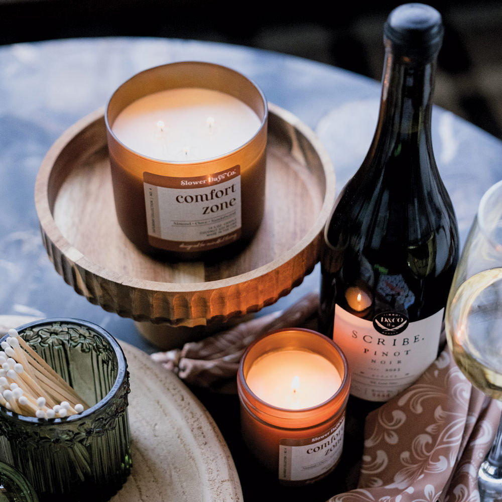A cozy evening lifestyle scene featuring Slower Days Co. Comfort Zone candles on a wooden pedestal tray alongside a bottle of Pinot Noir and a green glass match holder.