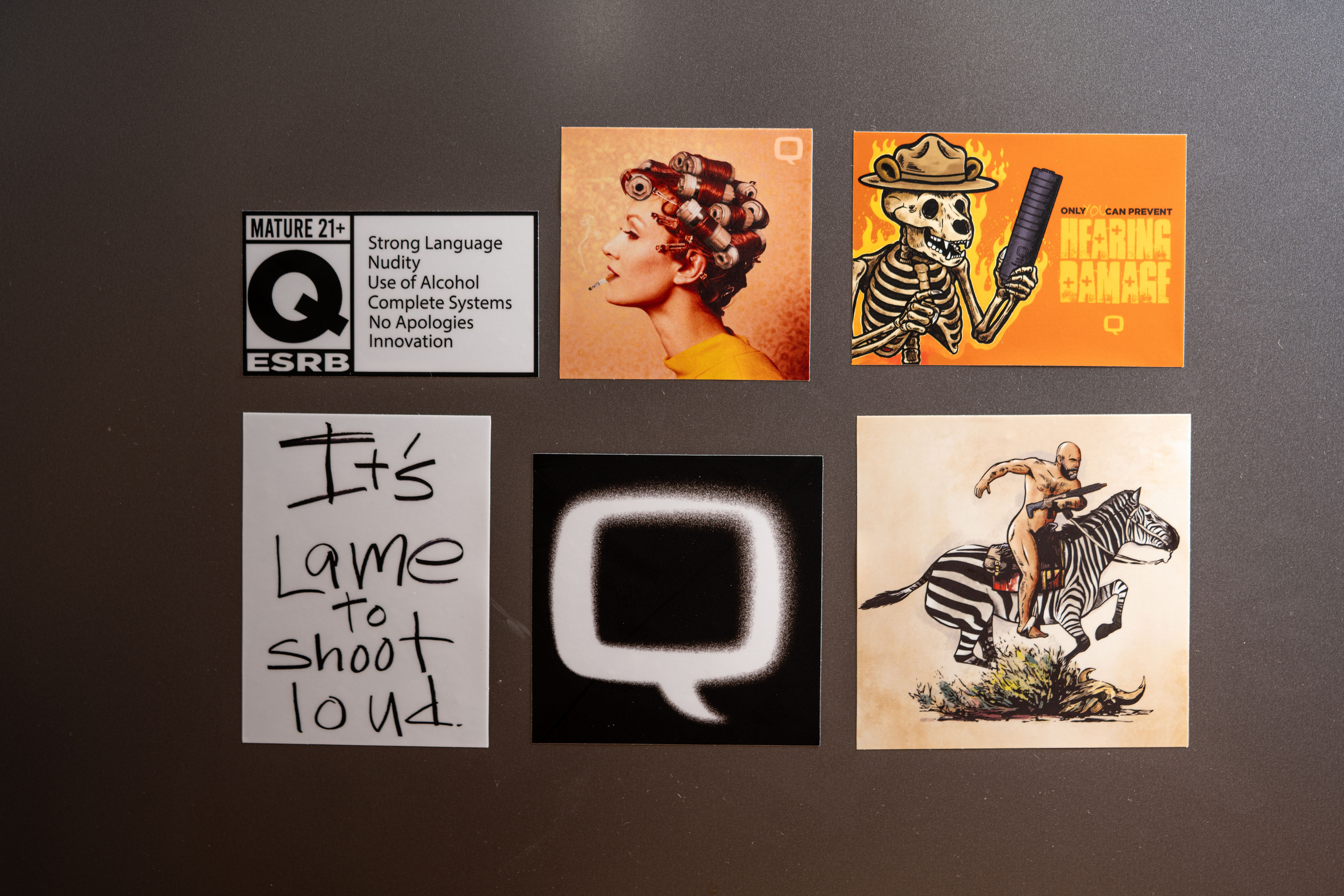 A close-up image of six unique Q-branded stickers arranged on a flat surface. Designs include a spoof ESRB label, a retro woman with suppressors as curlers, a flaming skeleton ranger holding a silencer, bold text reading “It’s lame to shoot loud,” a spray-painted Q logo, and a bald man riding a zebra with a rifle.