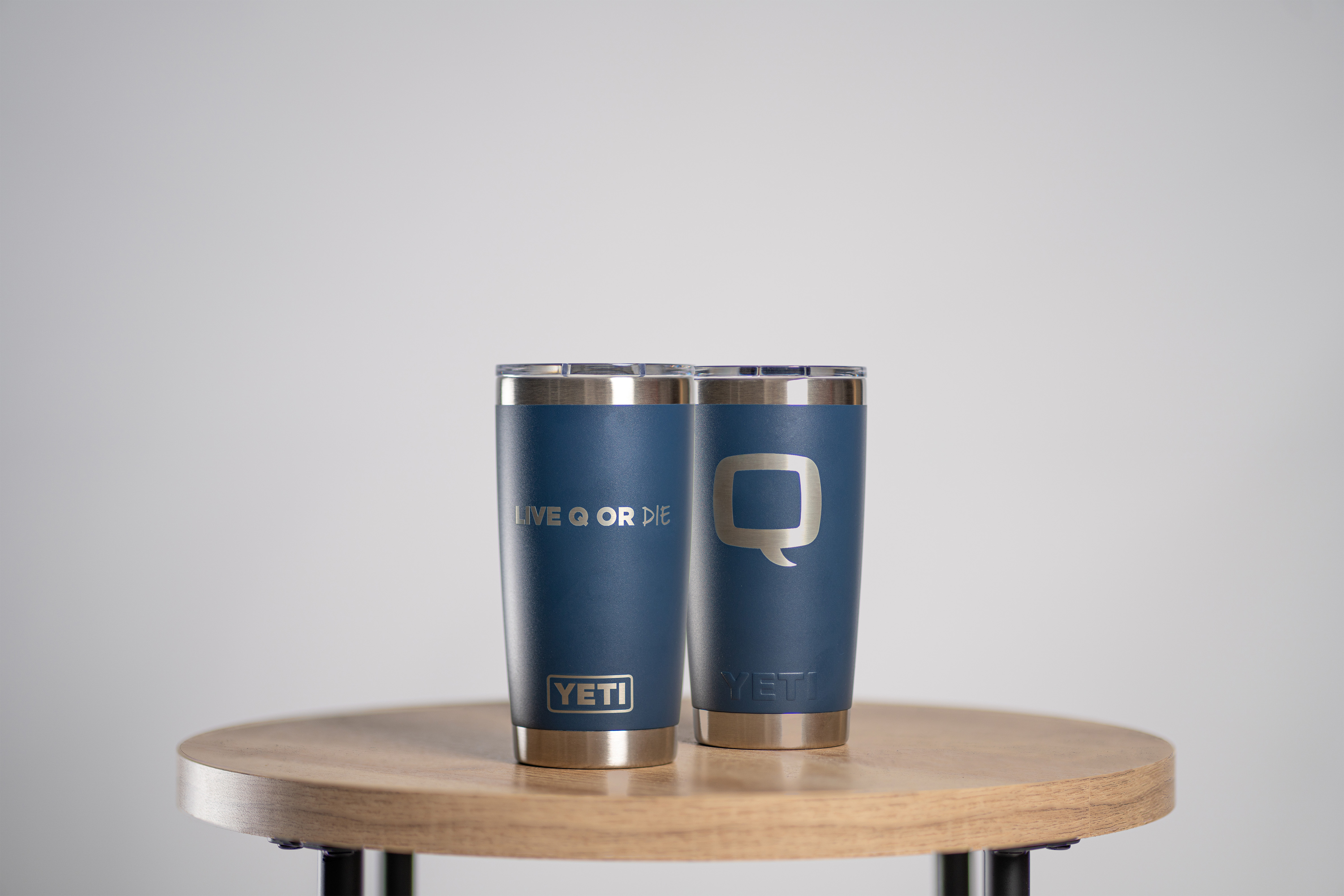 Two blue YETI tumblers on a wooden table, one with the Q logo and the other with the text ‘LIVE Q OR DIE.’