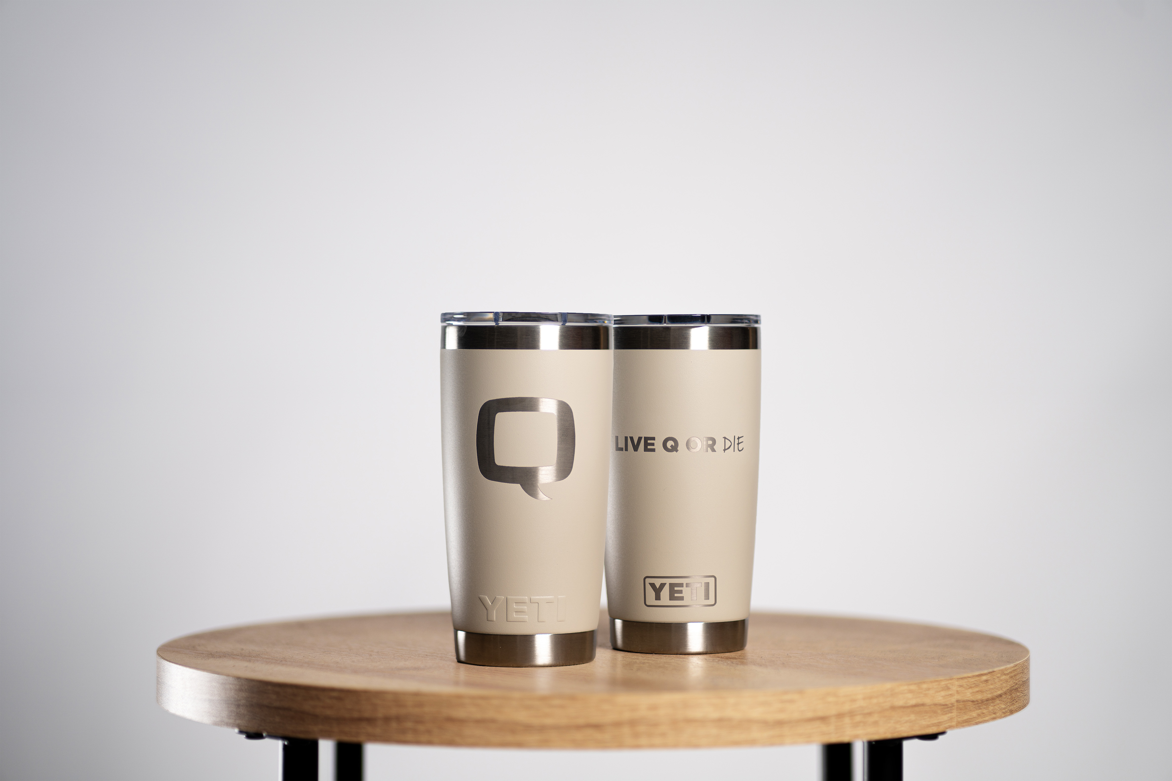 Two cream-colored YETI tumblers on a wooden table, one with the Q logo and the other with the text ‘LIVE Q OR DIE.’