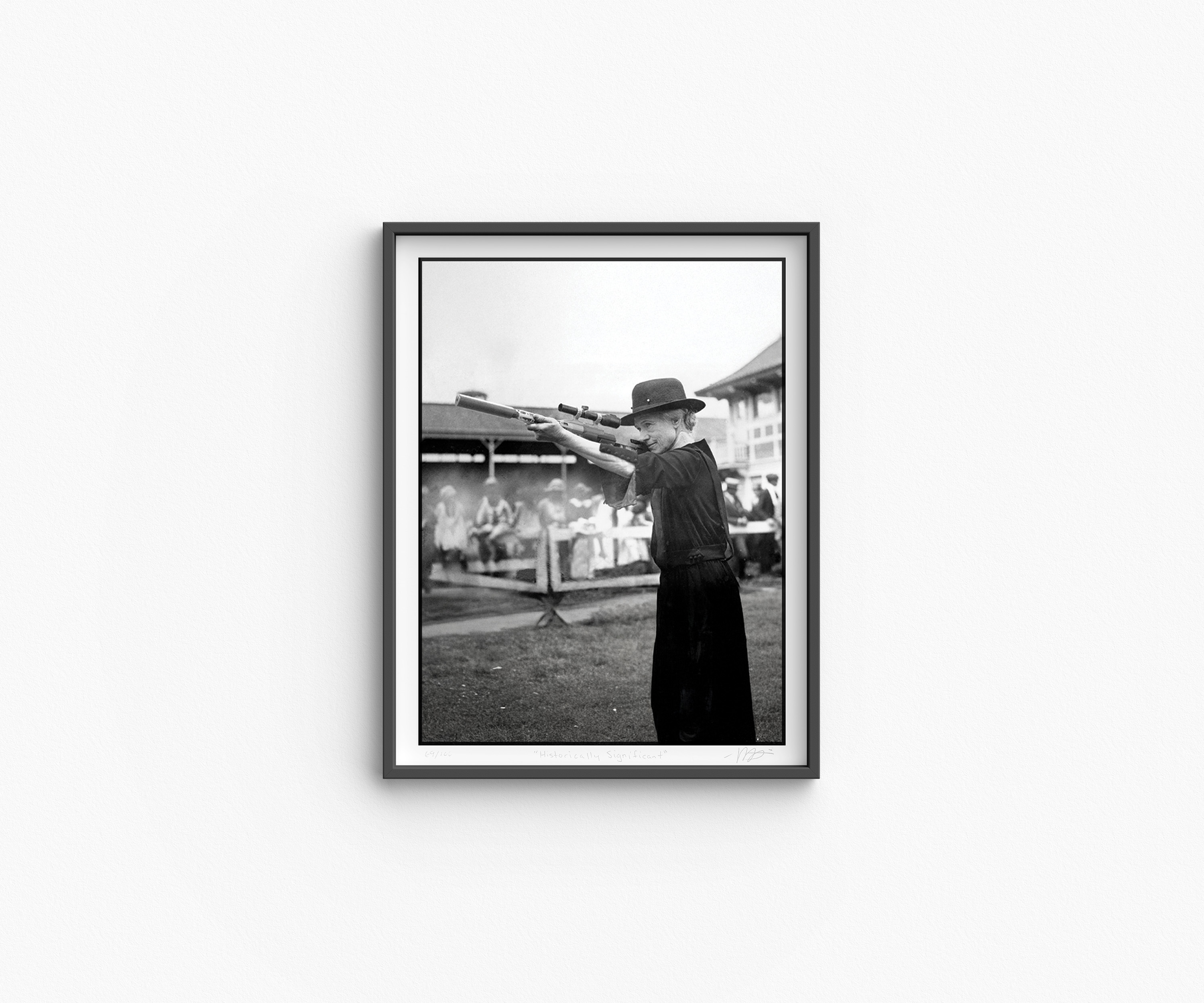 Framed black-and-white photograph of sharpshooter Annie Oakley aiming two rifles in opposite directions during a public exhibition. The crowd and fairground structures are visible in the background. The print is part of a limited edition series titled “Historically Significant.”