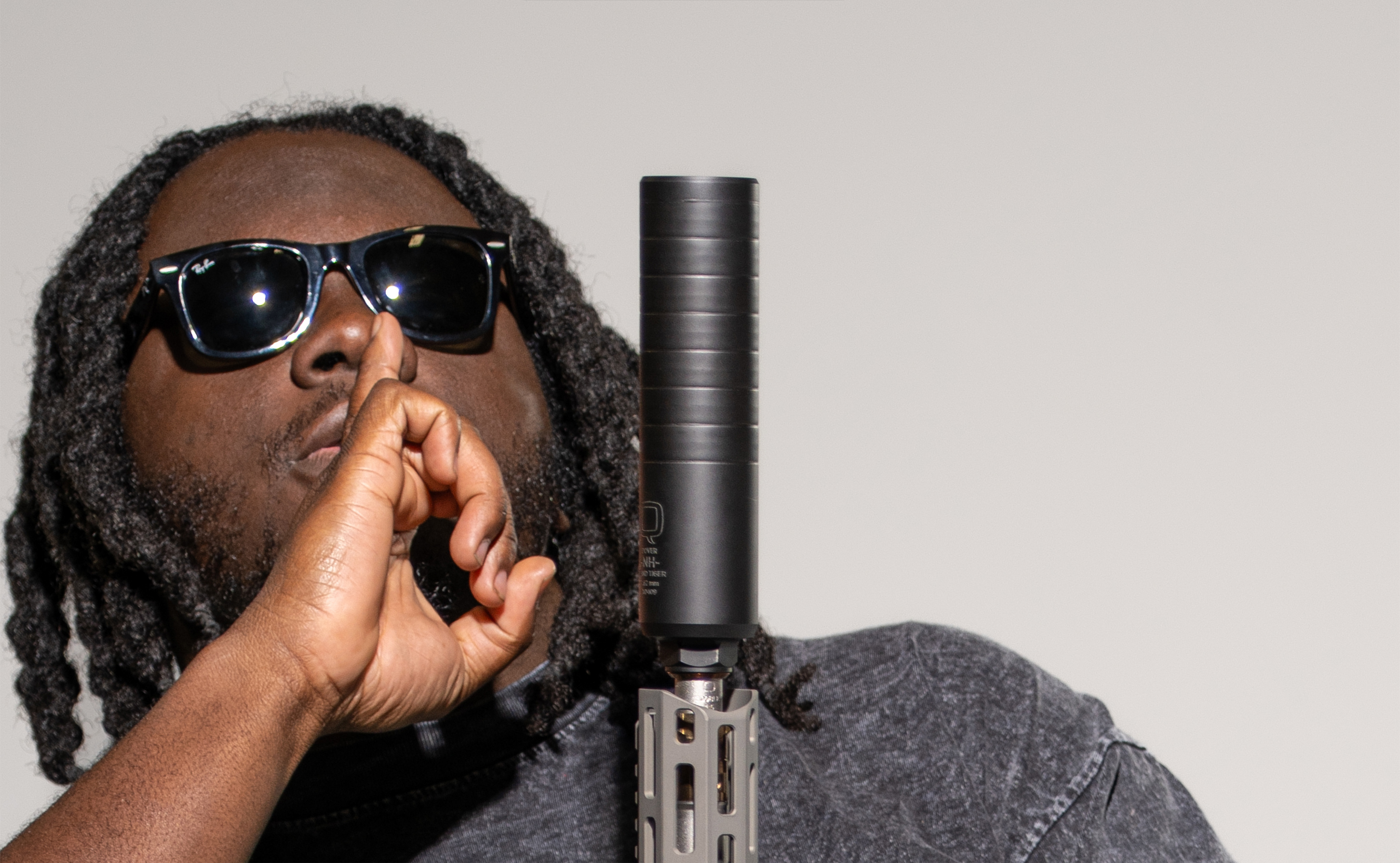 Man wearing sunglasses and a dark shirt holds a rifle vertically with a Blind Tiger suppressor attached. He gestures with a finger to his lips in a “shh” motion, emphasizing the silencer’s quiet performance. Clean, neutral background.