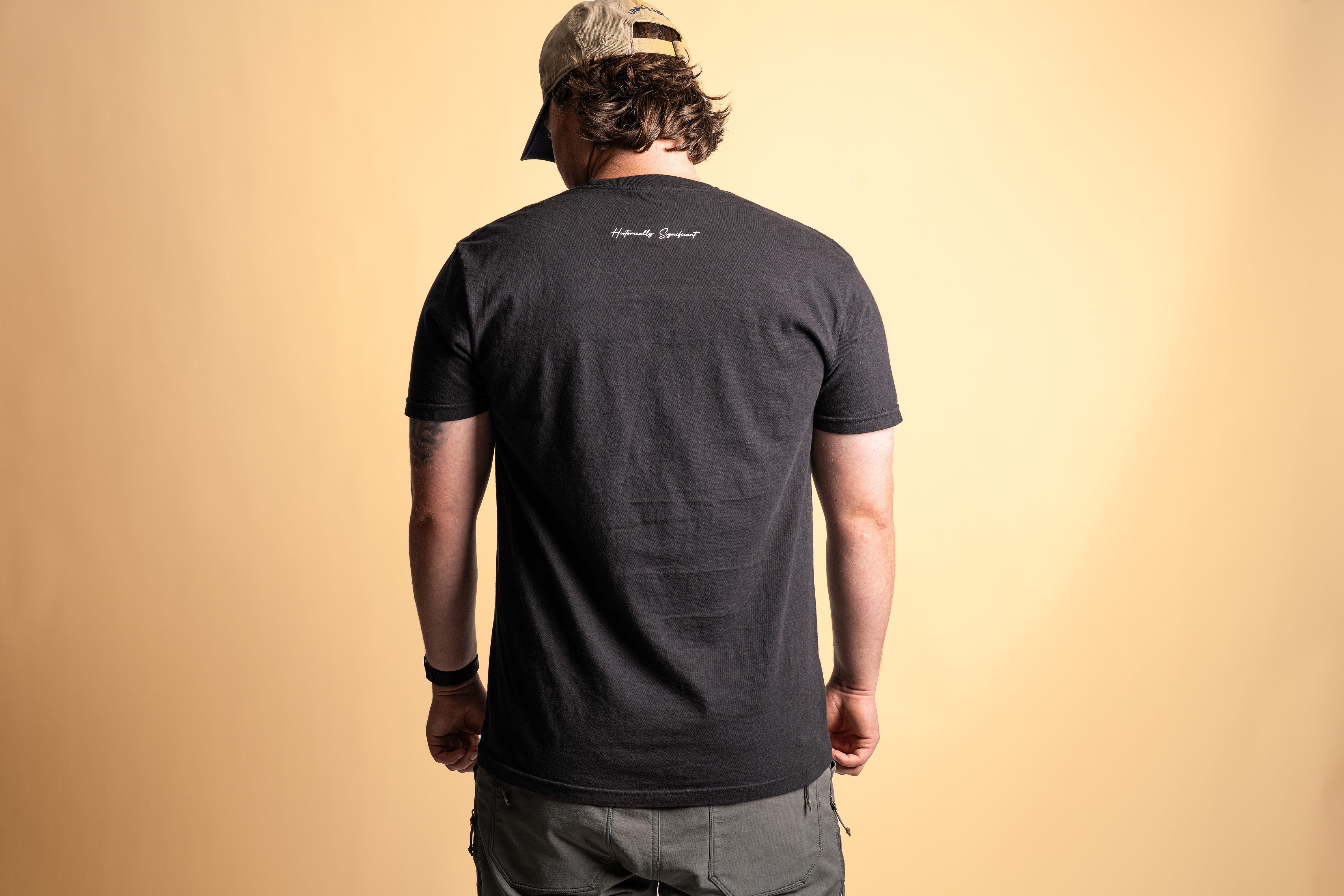 Back view of a man wearing a black t-shirt with “Historically Significant” printed at the neck; standing against a tan background with hands at his sides and head slightly turned.