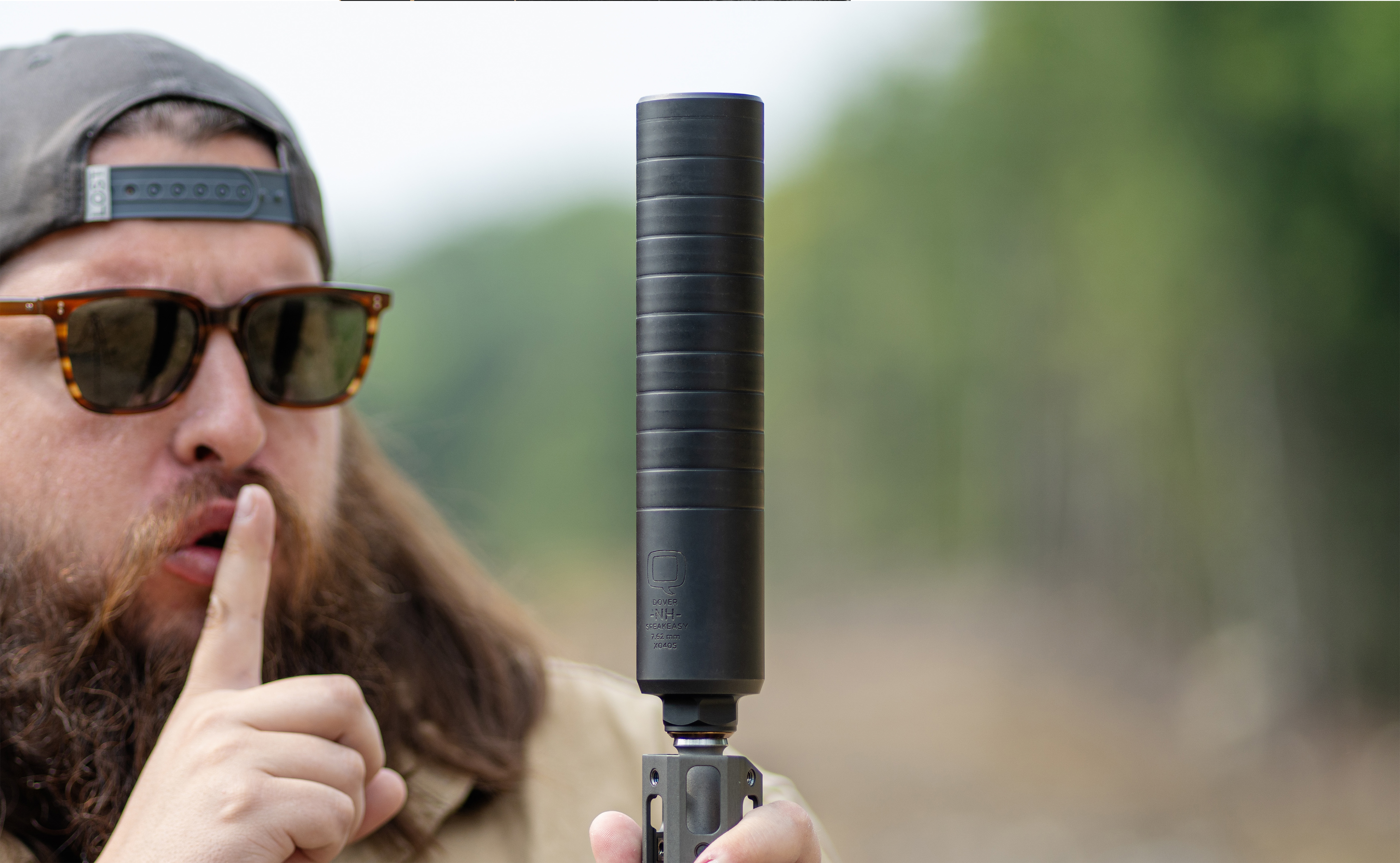 Man wearing sunglasses and a backwards cap holds a rifle upright with the Speakeasy suppressor attached. He gestures with a finger to his lips in a “shh” motion, emphasizing the suppressor’s quiet performance. The background is an outdoor wooded area with a soft blur.