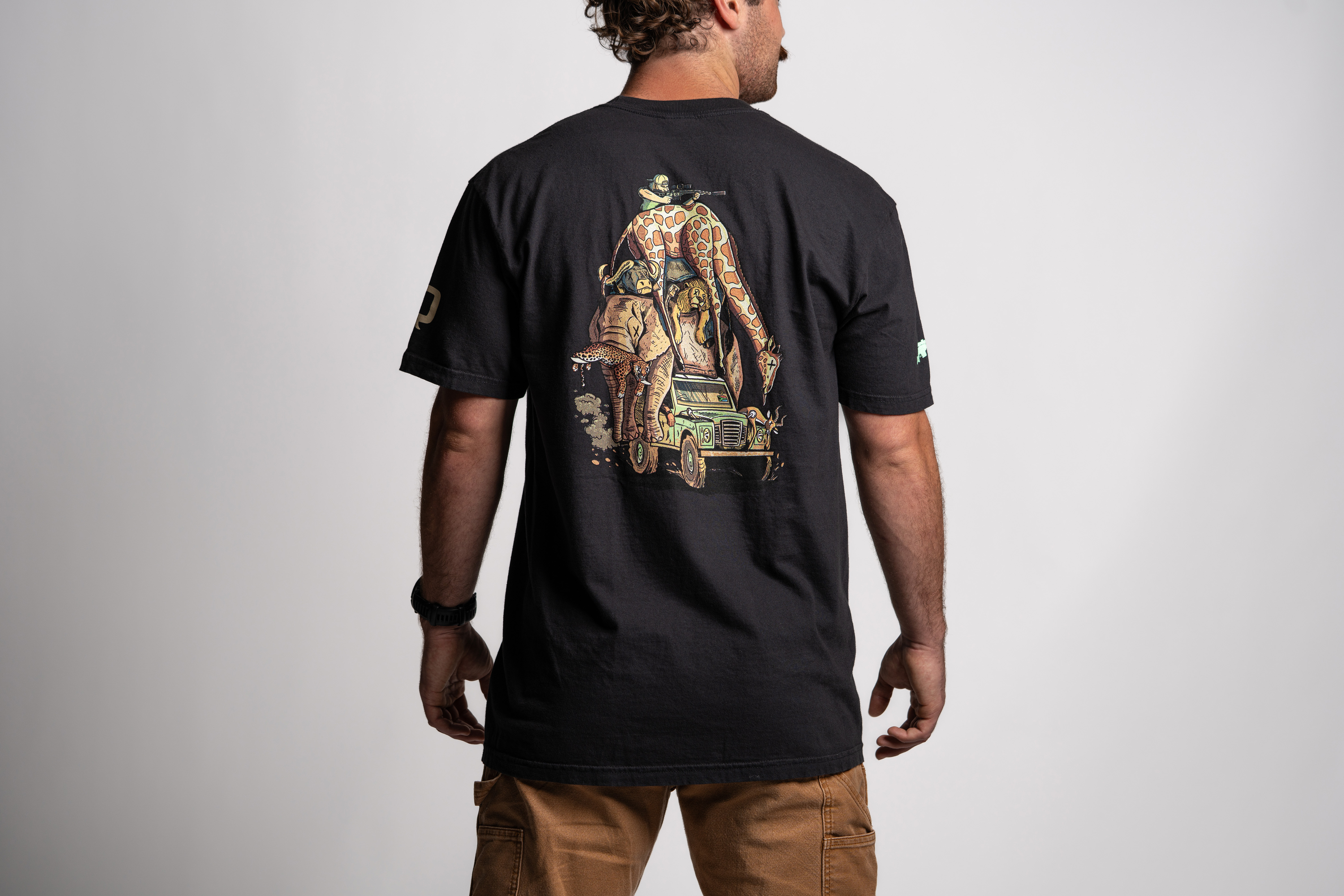 A man shown from the back wearing a black t-shirt featuring a large, colorful illustration. The graphic depicts a safari-style hunter with a rifle standing on top of a green off-road vehicle, which is overloaded with a stack of exotic animals including a giraffe, elephant, and lion. The man wears tan pants and stands against a plain light background.