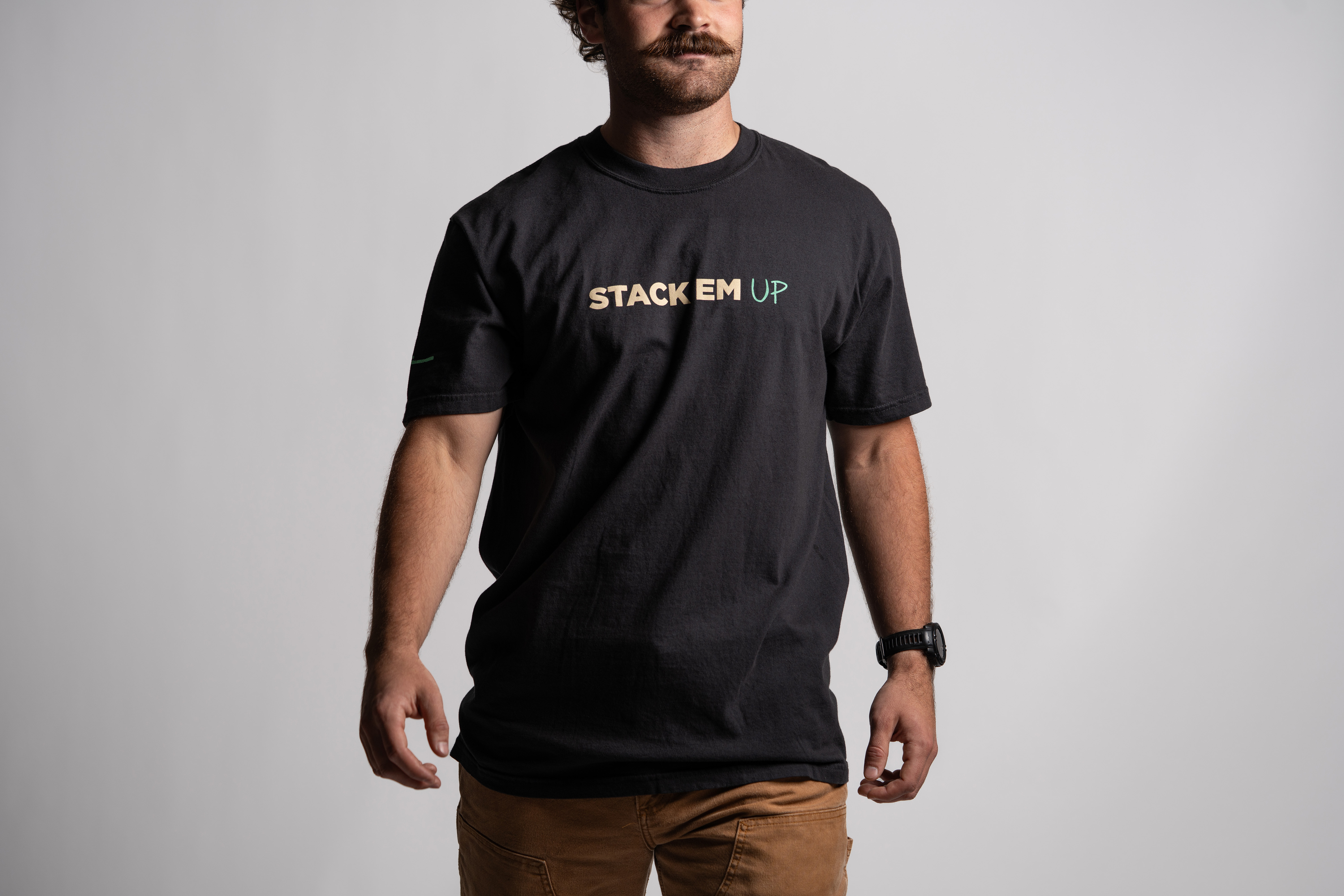 A man wearing a black t-shirt with the phrase “STACK EM UP” printed across the chest in tan and mint green letters. The model has a mustache and is standing against a plain light gray background, wearing tan pants and a black wristwatch.