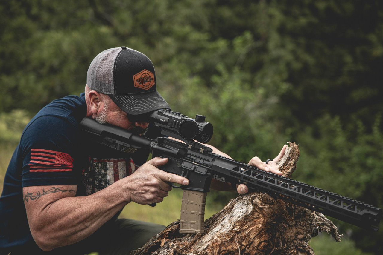 Shooter aiming a rifle equipped with a thermal scope while positioned behind a fallen tree outdoors.