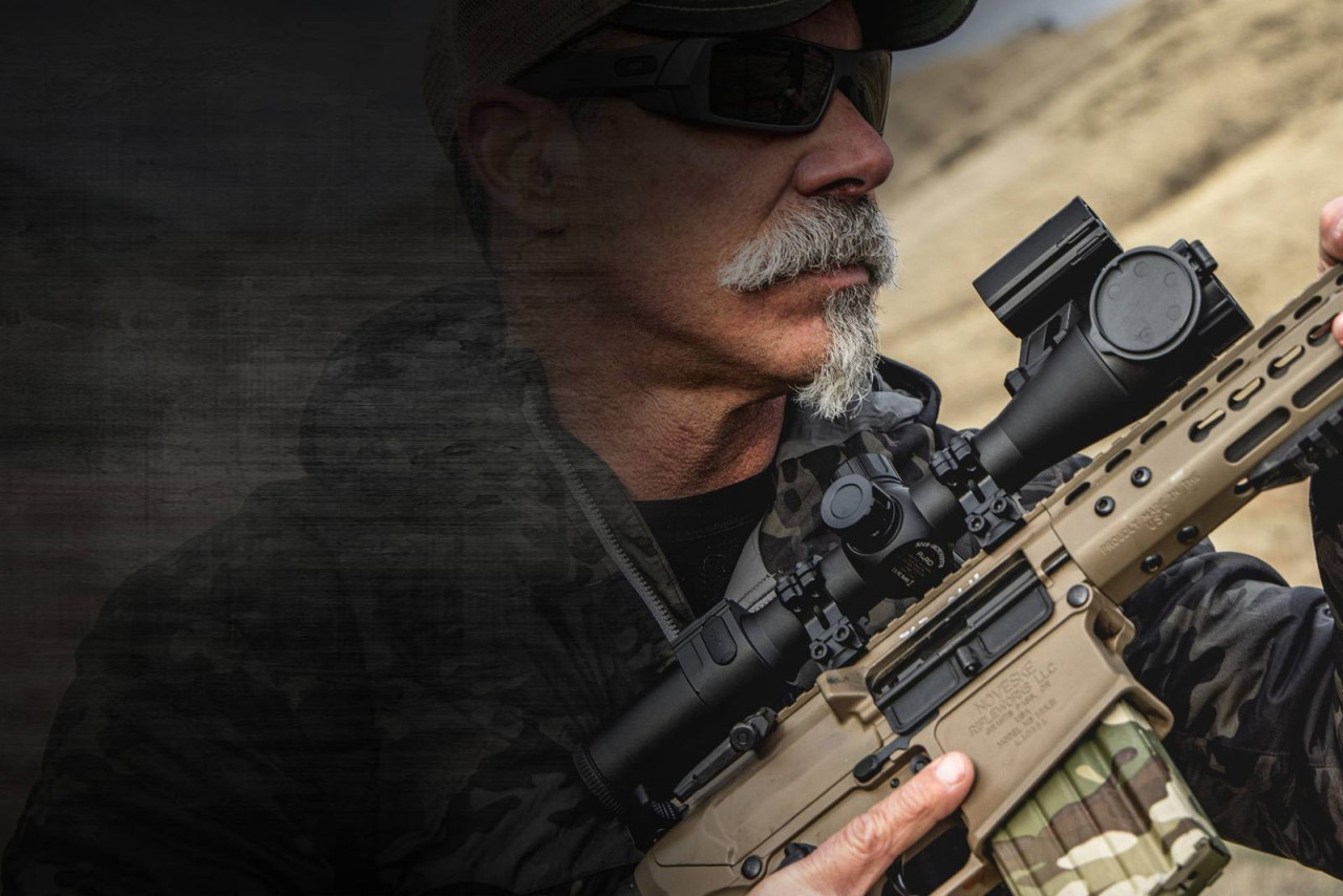 Shooter aiming a rifle fitted with a modern thermal optic, outdoors on a rugged terrain background.