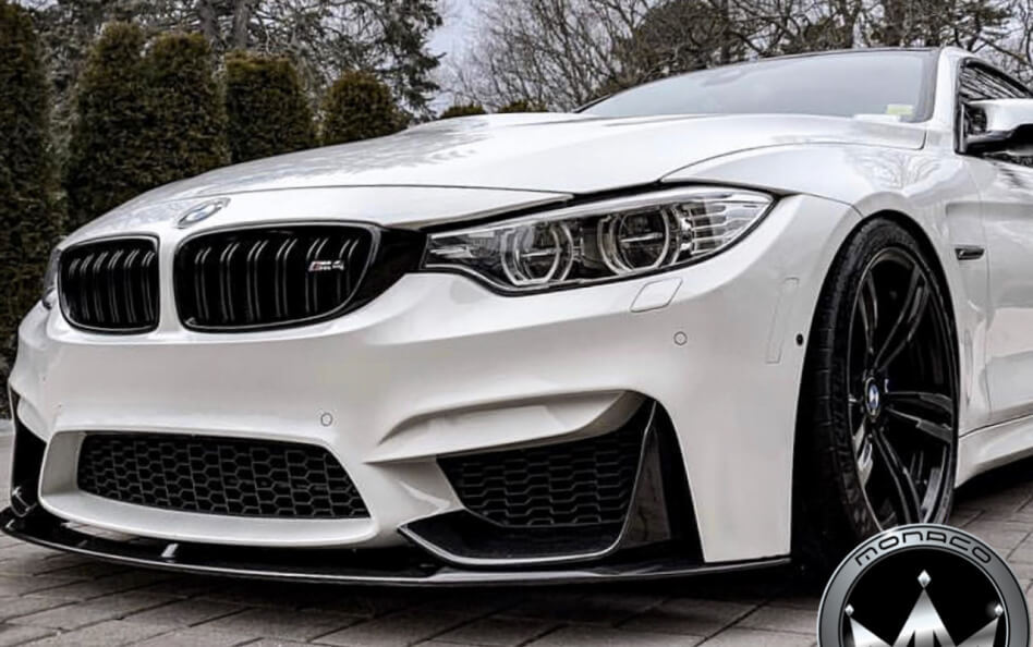 Front view of a white BMW M4 with aggressive styling upgrades, including blacked-out kidney grilles, a carbon fiber front lip, and black wheels.