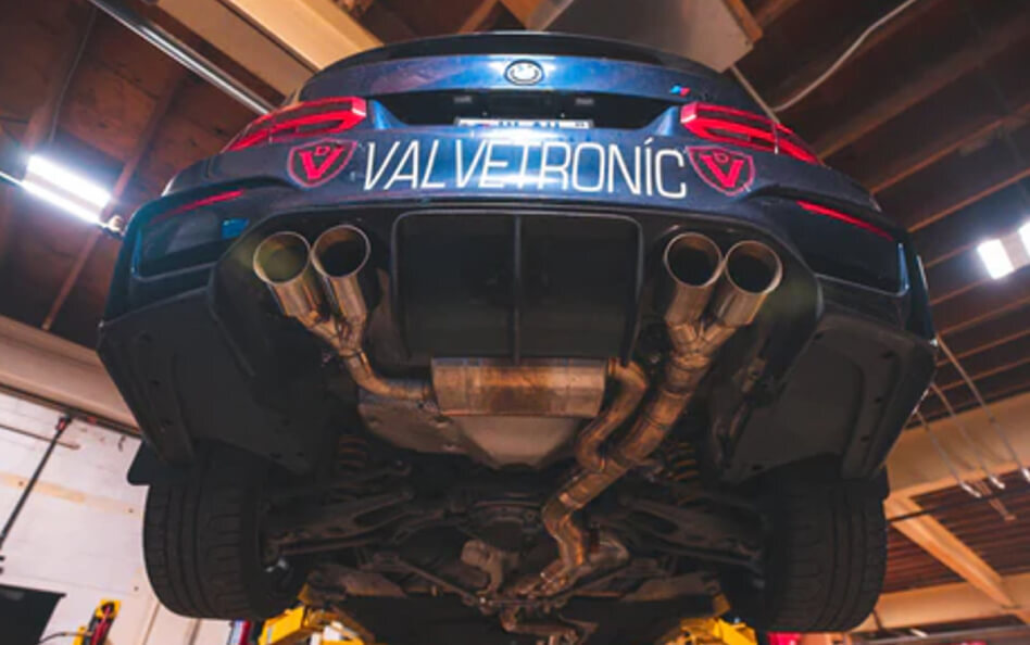 Underbody view of a sporty BMW with a custom quad-tip exhaust system labeled 'VALVETRONIC.' The car is lifted in a garage, showing the full aftermarket exhaust routing and components beneath the vehicle.