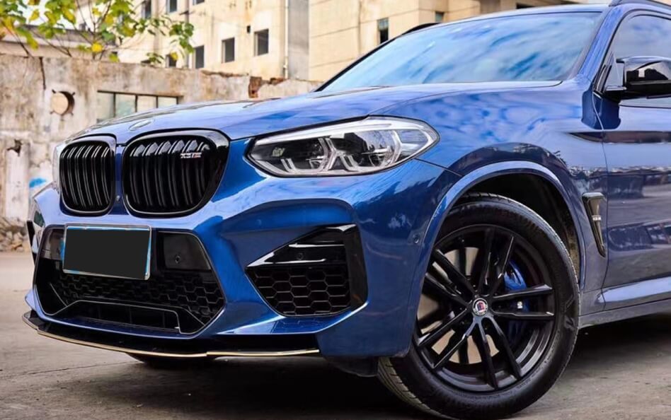 Front-side view of a blue BMW X3 M parked in an urban setting. The SUV features a blacked-out kidney grille, sporty front bumper with large air intakes, sleek LED headlights, and black multi-spoke alloy wheels with a performance brake system. The aggressive styling emphasizes the vehicle's high-performance capabilities.