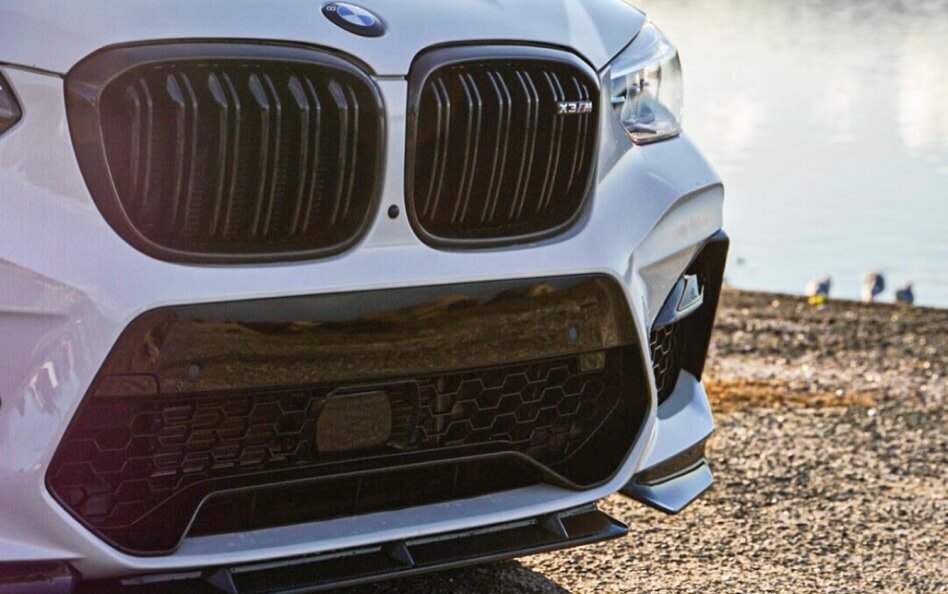 Front view of a white BMW X3 M bumper with blacked-out kidney grilles and lower mesh grille near a body of water.