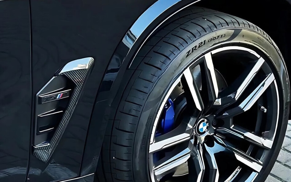 Close-up of a BMW M wheel with low-profile tire, blue brake caliper, and carbon fiber side vent detail.