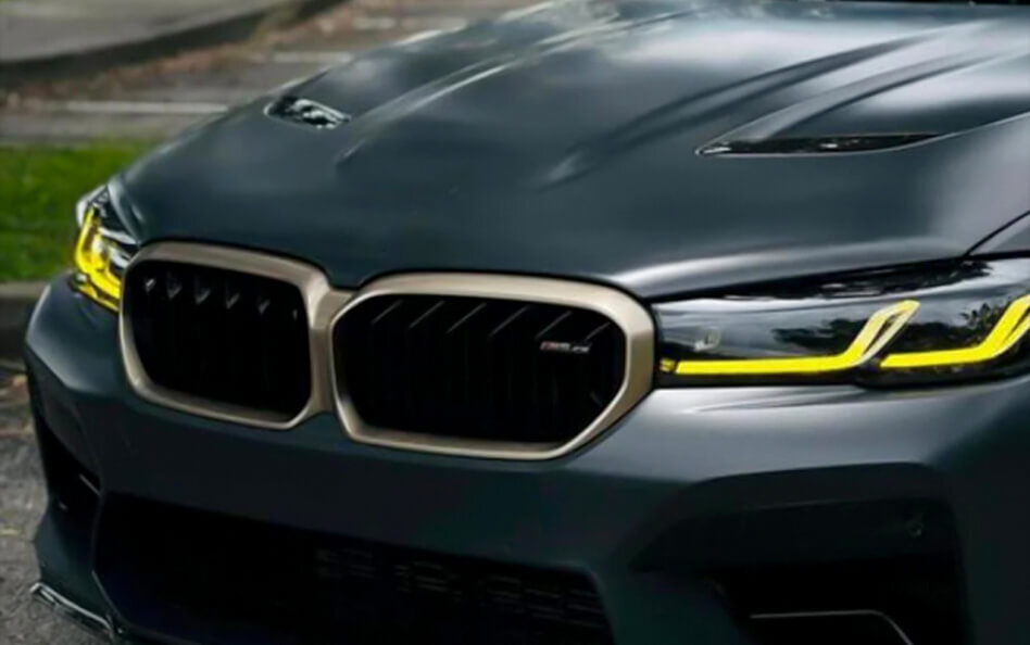 Front view of a matte dark gray BMW M5 CS showcasing a bold kidney grille with bronze trim, aggressive hood vents, and striking yellow LED daytime running lights.