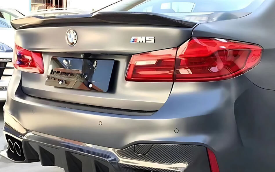 Rear view of a matte black BMW M5 featuring bold red LED tail lights, a carbon fiber trunk spoiler, and a quad exhaust system integrated into an aggressive rear diffuser. 