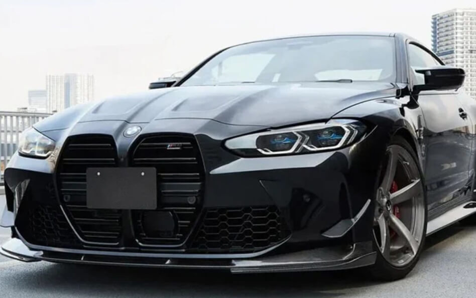 Front view of a sleek black BMW M4 with aggressive styling upgrades. The car features a large vertical kidney grille, sharp LED headlights with blue accents, a carbon fiber front lip spoiler, and large aftermarket wheels with red brake calipers.