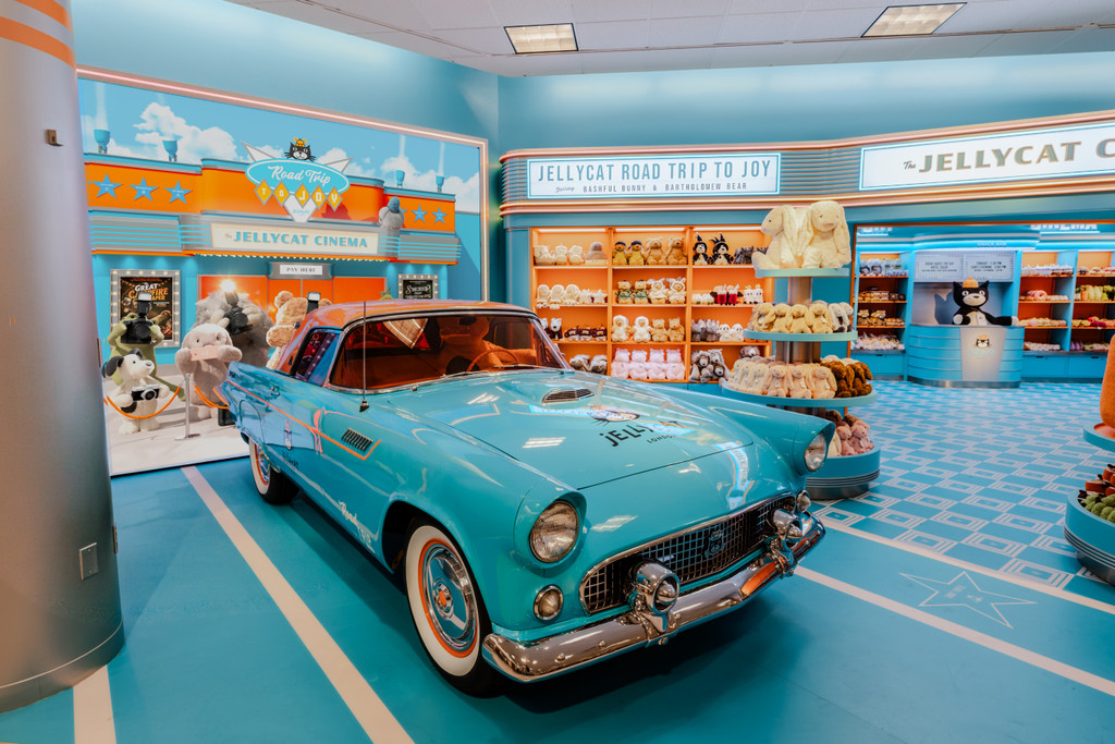 A vintage turquoise car is displayed inside a Jellycat Road Trip to Joy themed store featuring cinema-inspired décor, displays, and bright blue and orange interiors.